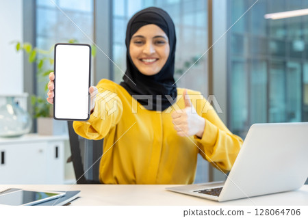 A smiling woman wearing a hijab shows a blank phone screen and gives a thumbs up, in an office setting. 128064706