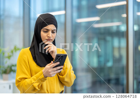 A woman wearing a black hijab and yellow shirt looks thoughtfully at her phone in a modern office setting. She has a hand on her chin. A woman wearing a black hijab and yellow shirt looks thoughtfully at her phone in a modern office setting. She has a hand on her chin. 128064718