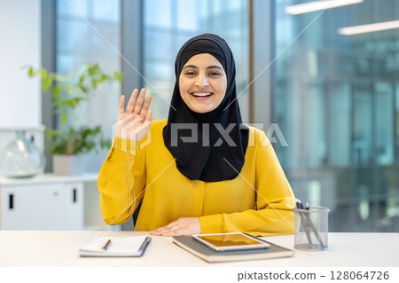 A smiling woman in a hijab waves at the camera, looking ready for a video call 128064726