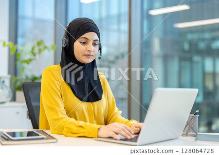 A woman wearing a hijab and headset works on a laptop in a modern office environment. A woman wearing a hijab and headset works on a laptop in a modern office environment. 128064728
