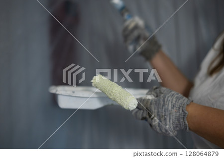 Close-up of a paint tray and roller in the hands of a painter. A professional painter dipped a small roller into a tray filled with white paint, preparing to continue painting a wall. 128064879