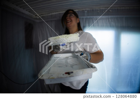 Woman painting a room with a roller brush, wearing protective gear. The space is covered in plastic sheeting, indicating a renovation or home improvement project in progress. 128064880