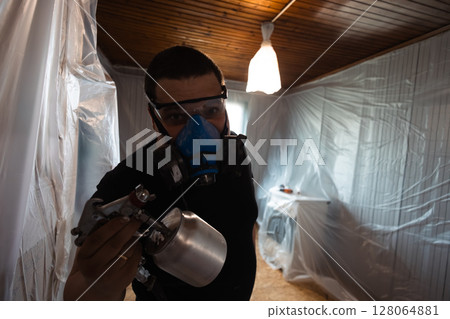 Painter wearing protective gear and using a spray gun for home renovation. The room is covered with plastic sheeting, indicating a focus on safety and cleanliness during the painting process. Painter wearing protective gear and using a spray gun for home renovation. The room is covered with plastic sheeting, indicating a focus on safety and cleanliness during the painting process. 128064881