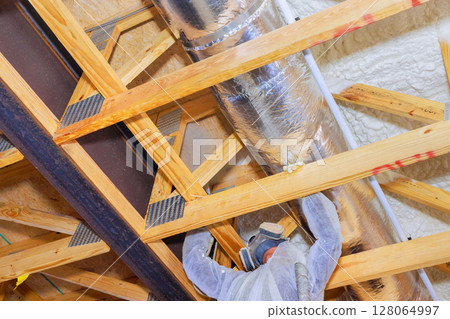 Construction worker is fitting thermal barrier insulation material above wooden ceiling frame in attic house being built. 128064997