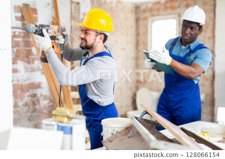 Builder drills a wall with a hammer drill, and his colleague makes notes Builder drills a wall with a hammer drill, and his colleague makes notes 128066154