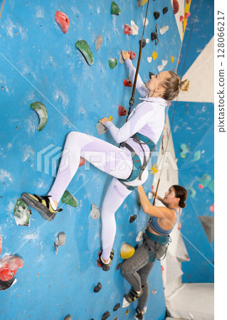 Focused woman climbing on bouldering wall with safety rope Focused woman climbing on bouldering wall with safety rope 128066217