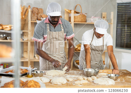 Man employee divide lump of dough by cutter, male apprentice rolling out dough with rolling pin 128066230
