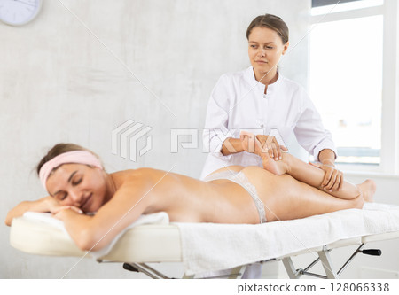 Female masseuse administering full-body sports massage to woman, stretching quadriceps 128066338