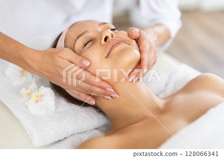 Young female client relaxing in spa salon during massage of face Young female client relaxing in spa salon during massage of face 128066341