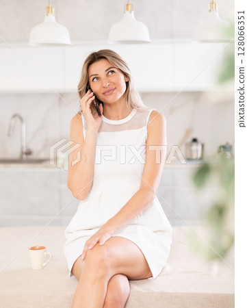Delightful blonde drinking coffee and talking on the phone while sitting on kitchen table 128066351