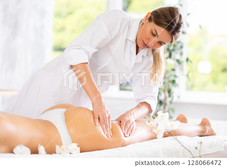 Professional young masseuse performing thigh and butt massage to female client Professional young masseuse performing thigh and butt massage to female client 128066472