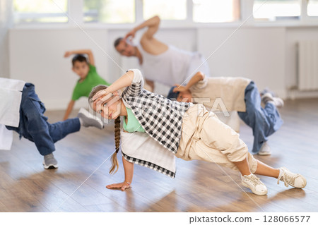 Smiling sports teen girl performing krump movements on floor with group children in breakdance studio with male coach Smiling sports teen girl performing krump movements on floor with group children in breakdance studio with male coach 128066577