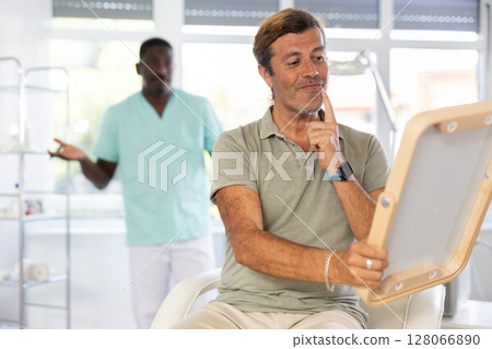 Man dissatisfied with results of rejuvenating at cosmetologist and meticulously examines reflection 128066890