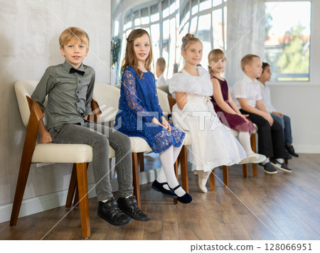 Little schoolchildren wearing elegant dresses sitting on chairs in school hall Little schoolchildren wearing elegant dresses sitting on chairs in school hall 128066951