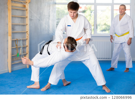 Two men practice grabbing and throwing sports mats during judo training under the guidance of an experienced mature coach 128066996
