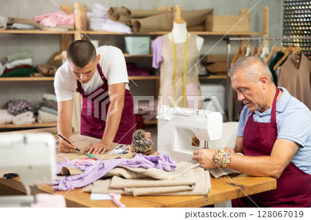 Mature male tailor sewing next to assistant who paints 128067019