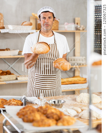 Caucasian male cook-baker worker with loafs of bread, shows many products Caucasian male cook-baker worker with loafs of bread, shows many products 128067325