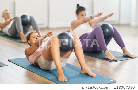 Young female Pilates instructor performing sit-ups with ball with group 128067350