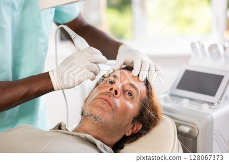 African male specialist uses ultrasonic import nozzle to care man client face skin 128067373