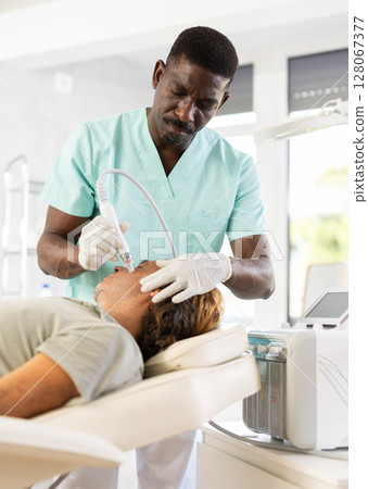 African male cosmetologist performs skin cleansing procedure for man client with beauty machine African male cosmetologist performs skin cleansing procedure for man client with beauty machine 128067377