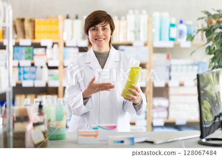 Friendly female pharmacist standing at drugstore with medical product in hands 128067484