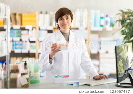 Friendly female pharmacist standing at drugstore with medical product in hands 128067487