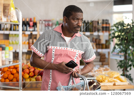 In store, African man with phone in hands looking for something on shelf of showcase In store, African man with phone in hands looking for something on shelf of showcase 128067569