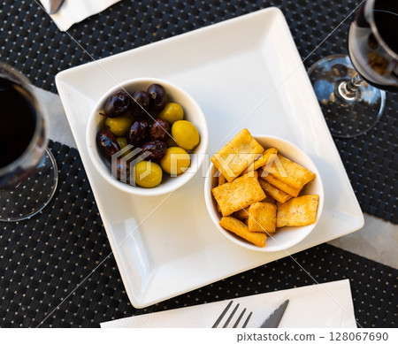 There are two bowls of snacks on white square plate - pickled olives and biscuits. There are two bowls of snacks on white square plate - pickled olives and biscuits. 128067690