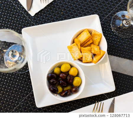 There are two bowls of snacks on white square plate - pickled olives and biscuits. 128067713