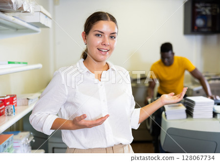 Happy young woman in white shirt sticking out her left hand to show the workflow in the printing house Happy young woman in white shirt sticking out her left hand to show the workflow in the printing house 128067734