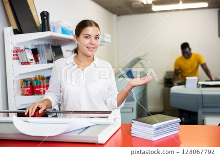 Cheerful young woman in white shirt smiling at the camera and using paper cutter in the printing house Cheerful young woman in white shirt smiling at the camera and using paper cutter in the printing house 128067792
