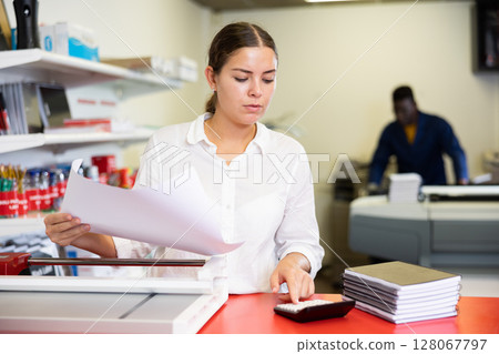 Concentrated young woman in white shirt using paper cutter on the table with planners and calculator in the printer house Concentrated young woman in white shirt using paper cutter on the table with planners and calculator in the printer house 128067797