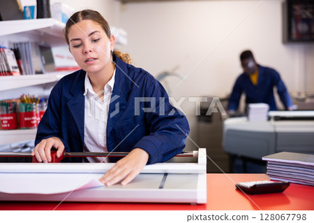 Serious young woman in a blue uniform using paper cutter on the table with planners and calculator in the printer house 128067798
