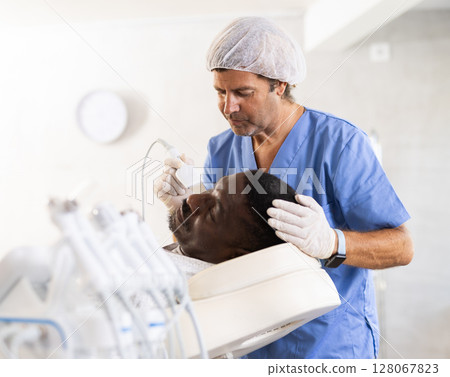 Cosmetologist performing facial cleansing with ultrasonic shovel on black man Cosmetologist performing facial cleansing with ultrasonic shovel on black man 128067823