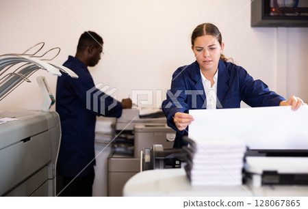 Serious young woman in uniform loading large format paper in a plotter in the print shop 128067865