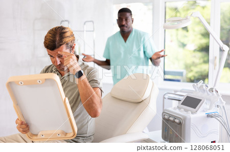 Man scrutinizing face in mirror after cosmetic procedures with confused cosmetologist 128068051