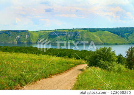 A rural field road leading to a calm river with hilly banks A rural field road leading to a calm river with hilly banks 128068250