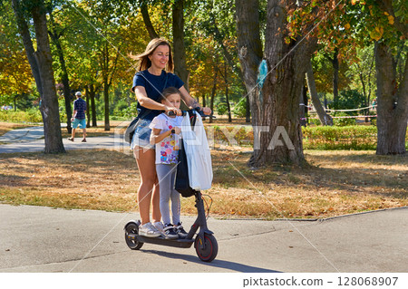 Cheerful mother and daughter riding electric scooter in summer park Cheerful mother and daughter riding electric scooter in summer park 128068907