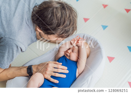 Loving father interacts with his 3-month-old baby lying in a cozy baby nest at home. Tender bonding moment, fatherhood, and early connection in a peaceful family setting 128069251