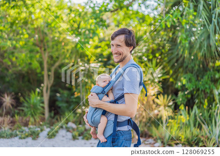 Young father walks in nature with his baby son nestled close in a parent-facing ergonomic baby carrier. Bonding, warmth, and modern parenting concept in a peaceful outdoor setting 128069255