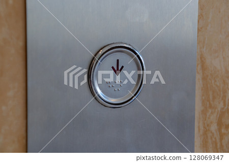 Close up view of an elevator button with downward arrow and braille dots on metal panel. 128069347