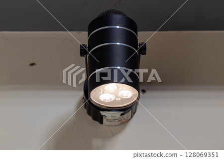 Black cylindrical LED spotlight fixture mounted on a wall emitting bright white light 128069351