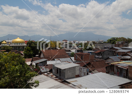 Golden dome of a mosque rises above traditional houses and trees with mountains in the background 128069374