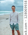 Handsome man in shirt relax on the beach. Laughing beach guy in shorts and shirt in cloudy day on the coast 128069938