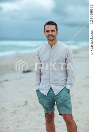 Handsome man in shirt relax on the beach. Laughing beach guy in shorts and shirt in cloudy day on the coast 128069938