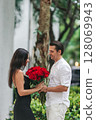 Young man gives his girlfriend a bouquet of red roses on Valentine's Day. The girl is wearing an elegant black dress. Valentine's Day Flowers 128069943