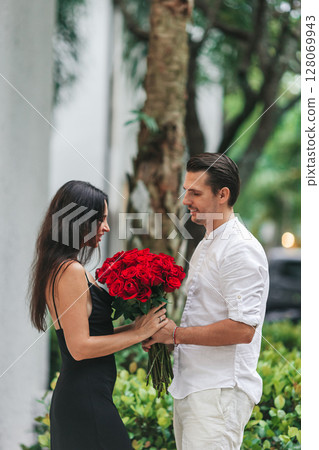 Young man gives his girlfriend a bouquet of red roses on Valentine's Day. The girl is wearing an elegant black dress. Valentine's Day Flowers 128069943