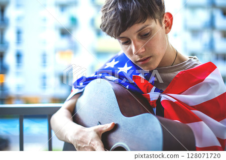 Serious teenage boy with American flag on shoulders playing guitar while sitting on balcony  128071720