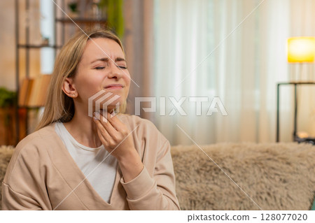 Adult woman touching throat at home, showing sore pain and discomfort while swallowing on home sofa Adult woman touching throat at home, showing sore pain and discomfort while swallowing on home sofa 128077020
