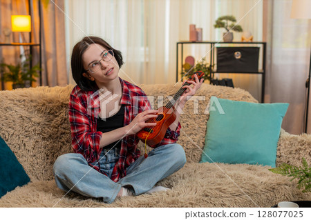 Young woman playing ukulele at home, focused on learning chord and smiling with satisfaction and joy Young woman playing ukulele at home, focused on learning chord and smiling with satisfaction and joy 128077025